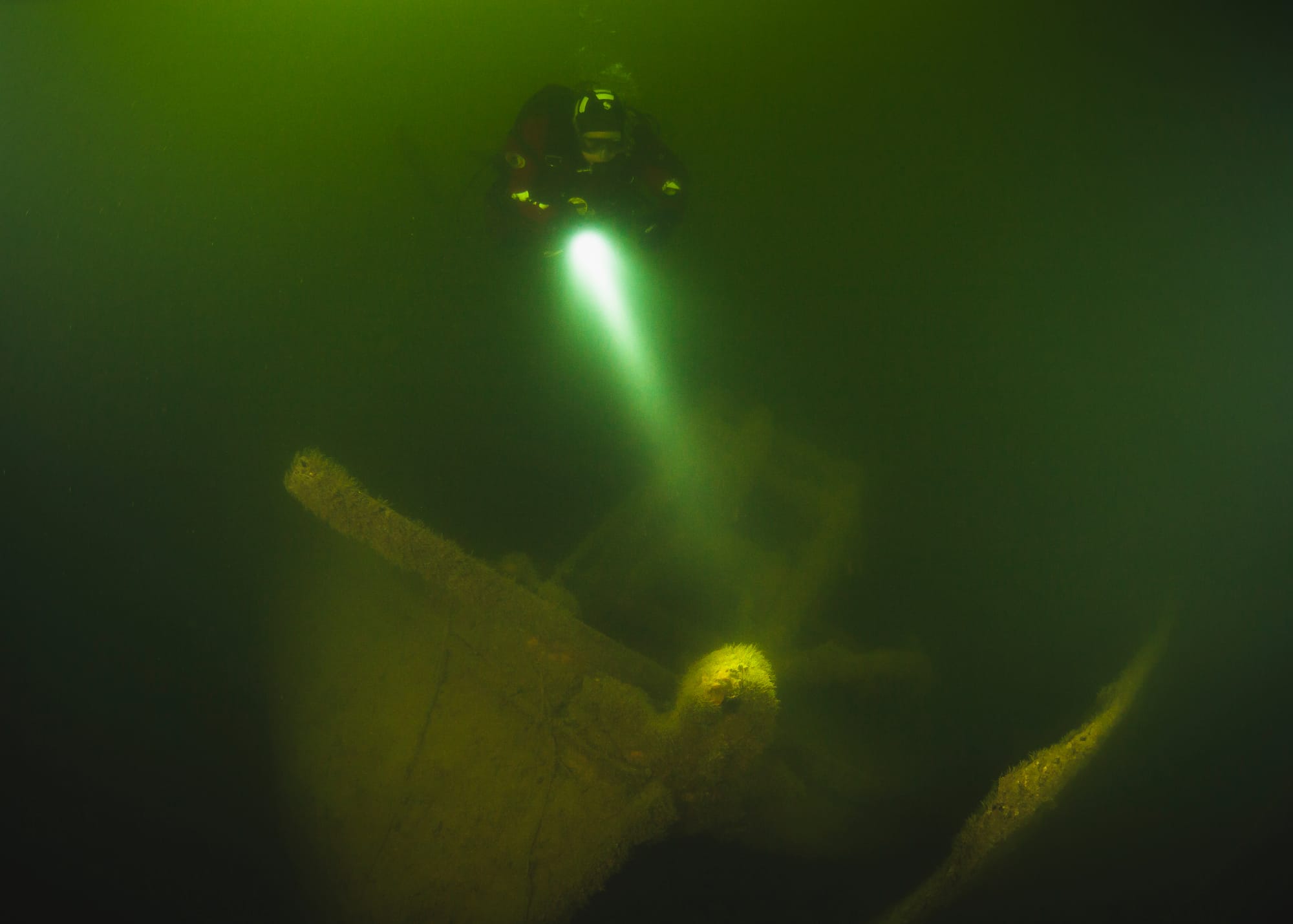 A diver in murky water, floating above a wreck and using a torch to see better